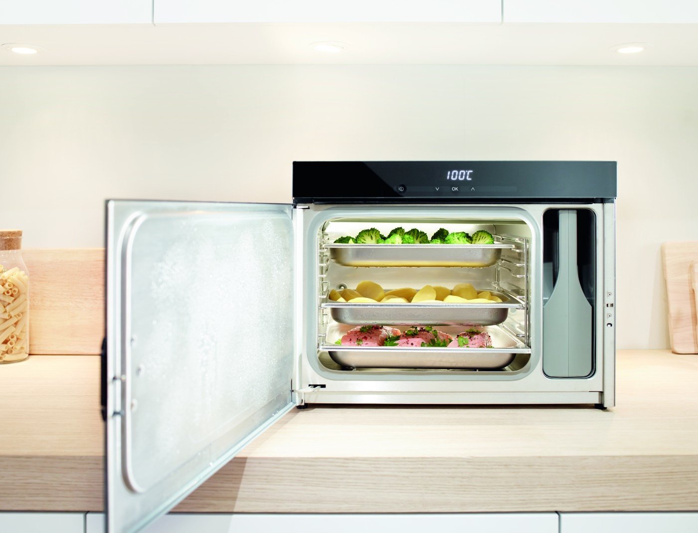 Le four à vapeur MIELE DG6019NR est posé, porte ouverte, sur le plan de travail d'une cuisine moderne ; à l'intérieur se trouvent des plateaux de brocolis, de pommes de terre et de poulet assaisonné.