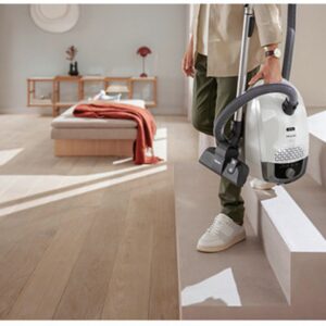 Une personne tenant un aspirateur Miele GUARD S1 BLEU NORDIQUE FLEX blanc, debout sur les marches d'un escalier dans un salon lumineux et moderne, doté d'un parquet.