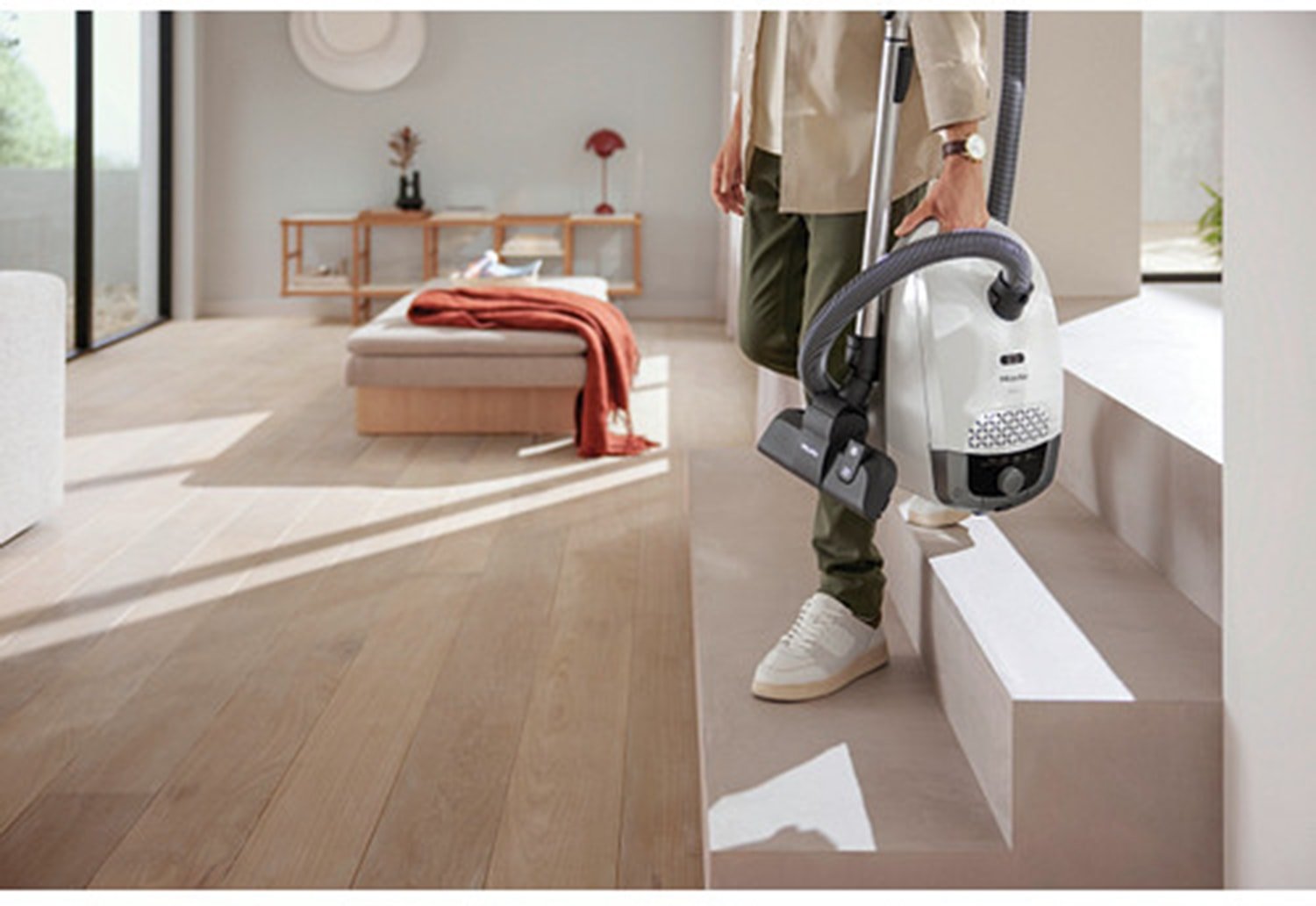 Une personne tenant un aspirateur Miele GUARD S1 BLEU NORDIQUE FLEX blanc, debout sur les marches d'un escalier dans un salon lumineux et moderne, doté d'un parquet.