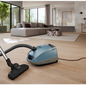 Un aspirateur-traîneau Miele GUARD S1 BLEU NORDIQUE FLEX est posé sur un parquet dans un salon moderne et lumineux.