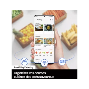 Une main tient un smartphone sur lequel s'affiche une application de cuisine, avec des plats et des légumes frais en arrière-plan, tandis que le réfrigérateur SAMSUNG RB50DG601ES9 se trouve à proximité, garantissant que les ingrédients restent frais et prêts à être utilisés pour chaque repas.