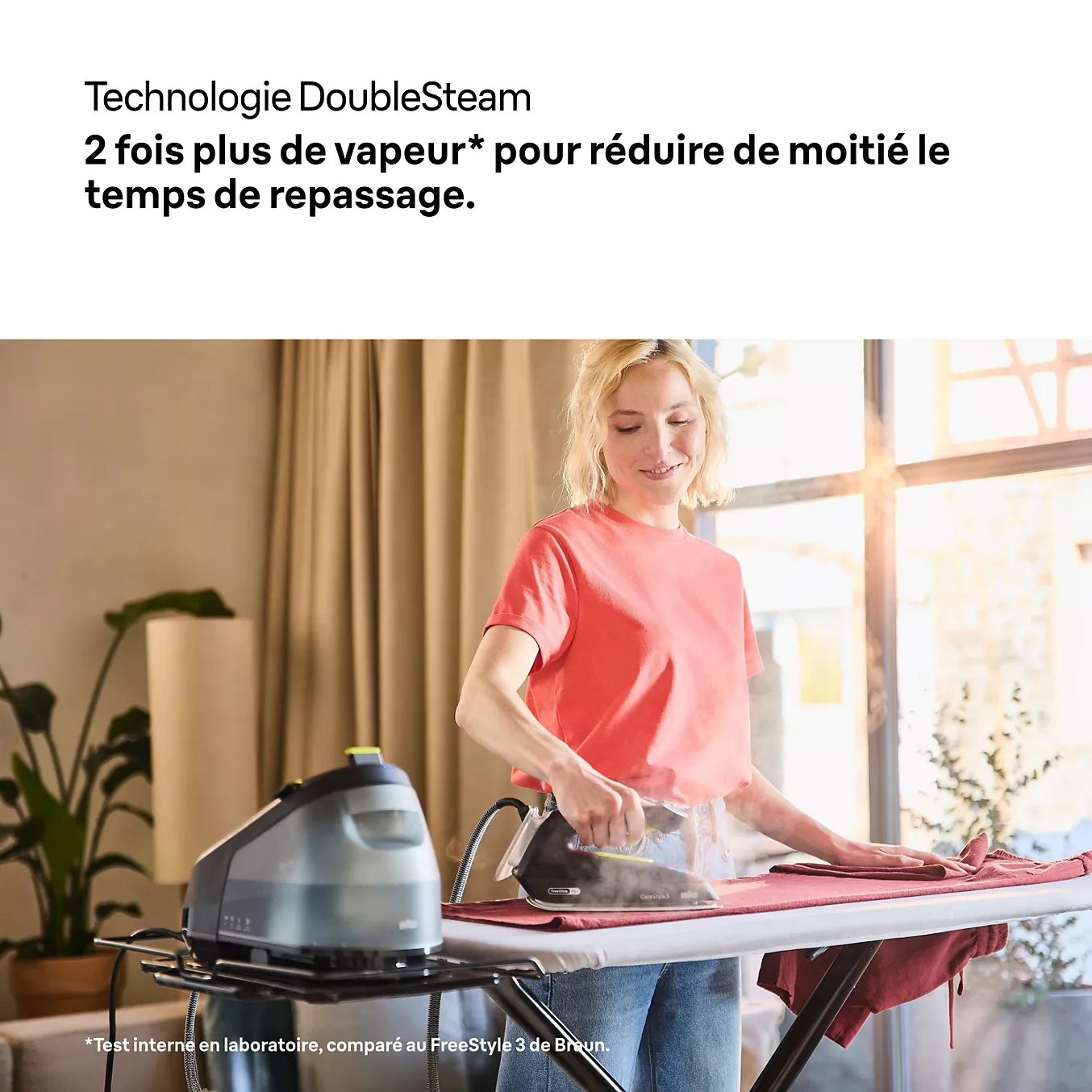 Une femme repasse une chemise chez elle, la lumière du soleil inondant la pièce à travers les grandes fenêtres derrière elle, et obtient un résultat parfaitement lisse grâce au fer à vapeur BRAUN IS3257BK, un appareil fiable.