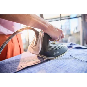 Une main repassant une chemise bleue à l'aide d'un fer à vapeur BRAUN IS3257BK, tandis que la lumière du soleil pénètre par la fenêtre en arrière-plan.