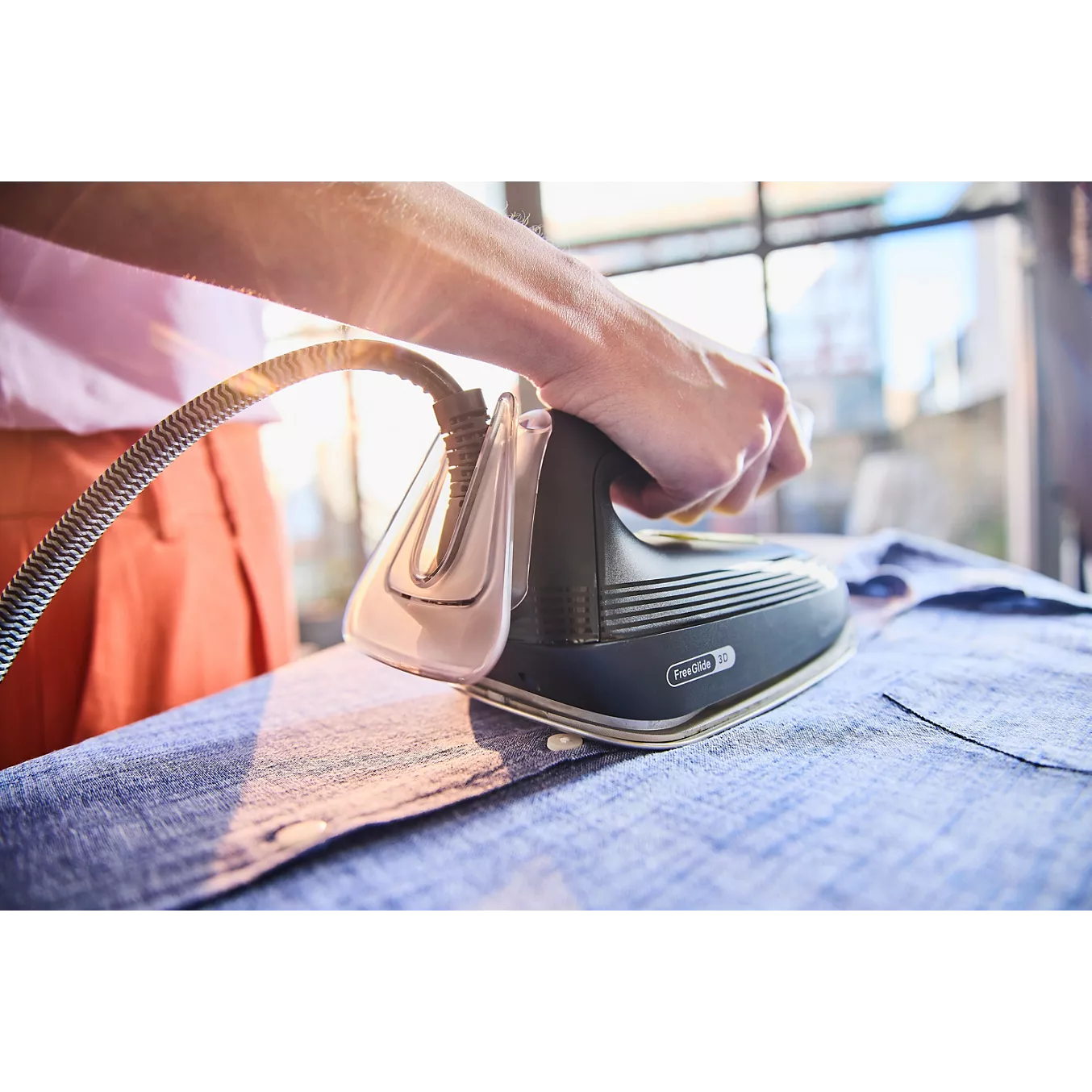 Une main repassant une chemise bleue à l'aide d'un fer à vapeur BRAUN IS3257BK, tandis que la lumière du soleil pénètre par la fenêtre en arrière-plan.