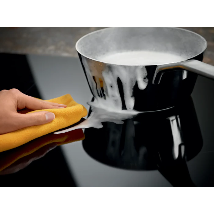 Une casserole déborde sur la cuisinière ; tandis qu'une main essuie le liquide renversé à l'aide d'un chiffon jaune, le lave-vaisselle ELECTROLUX LIS62351, situé à proximité, est prêt à faciliter le nettoyage.