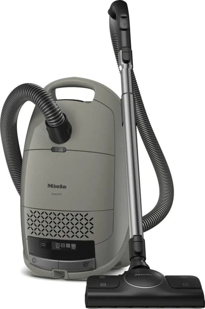 Aspirateur-traîneau Miele GUARD M1 SILENCE gris, équipé d'un tuyau, d'une poignée et d'un embout pour sol, se range à la verticale pour un nettoyage silencieux et efficace.