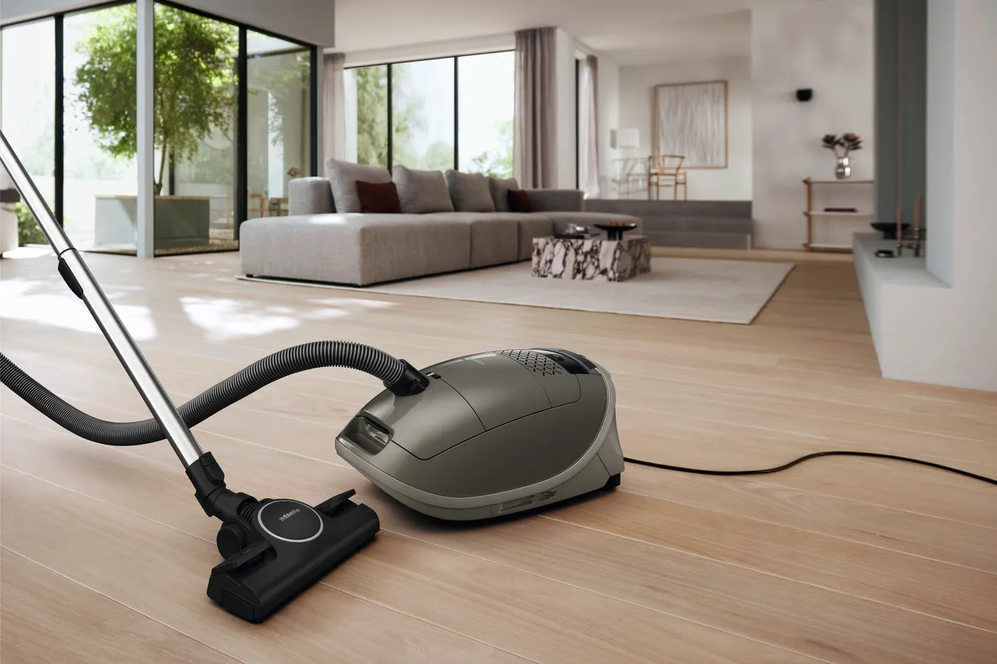 L'aspirateur Miele GUARD M1 SILENCE, doté de la technologie SILENCE, est posé sur un parquet dans un salon moderne agrémenté d'un canapé gris et de grandes fenêtres.