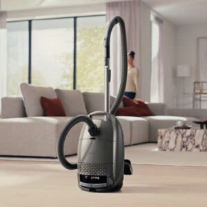 Un aspirateur Miele GUARD M1 SILENCE trône sur un parquet clair dans un salon moderne, avec en arrière-plan un canapé et de grandes fenêtres.