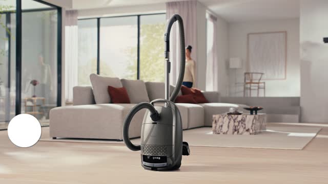 Un aspirateur Miele GUARD M1 SILENCE trône sur un parquet clair dans un salon moderne, avec en arrière-plan un canapé et de grandes fenêtres.