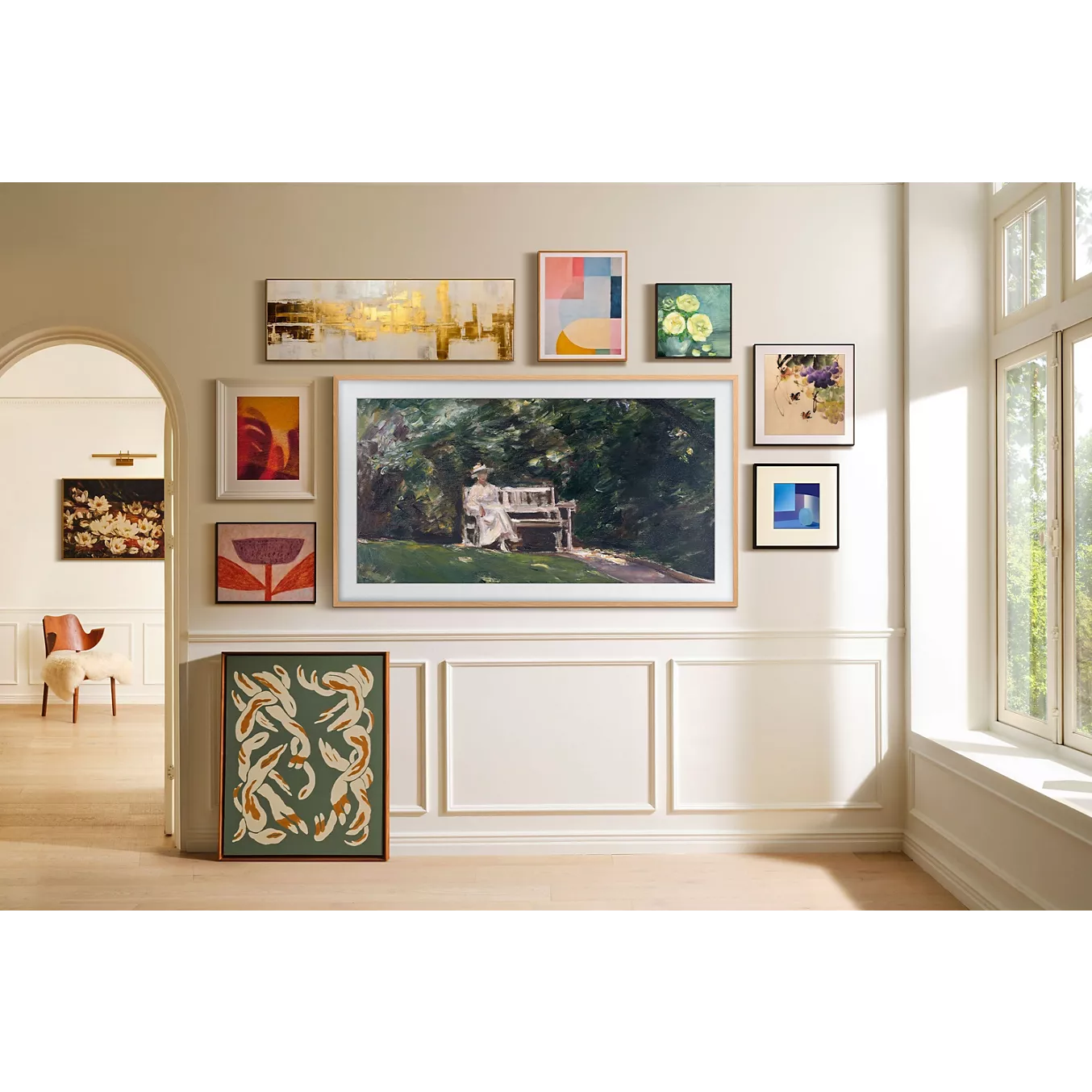 Un mur-galerie présentant une série d'œuvres d'art encadrées, dont l'élégant SAMSUNG TQ55LS03HW THE FRAME, dans une pièce lumineuse dotée de grandes baies vitrées et de lambris blancs.