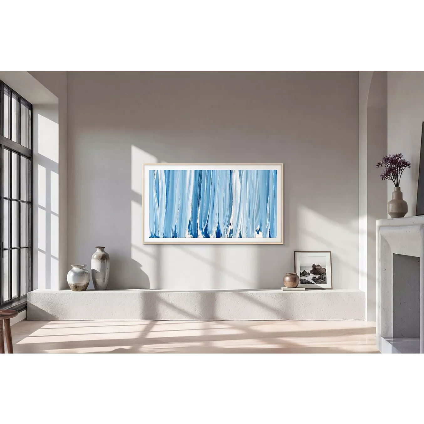 Dans un salon moderne, où trônent un tableau abstrait bleu, des poteries et des photos encadrées posées sur un rebord baigné de soleil, le téléviseur SAMSUNG TQ55LS03HW THE FRAME allie avec élégance technologie et art.