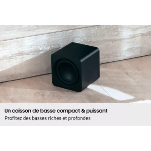 Le SAMSUNG HW-Q930H, un caisson de basses compact noir issu d'un système home cinéma, est posé sur un sol de couleur claire, près d'un mur, avec un texte en français affiché en dessous.